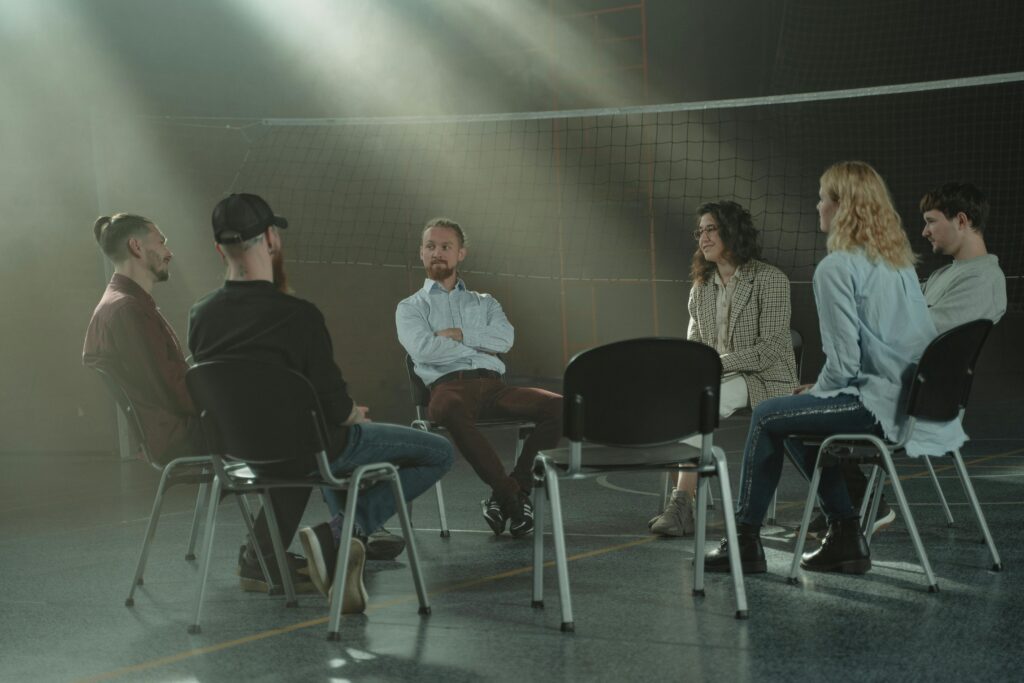 A group therapy session with six adults seated in a circle, discussing support and mental health.