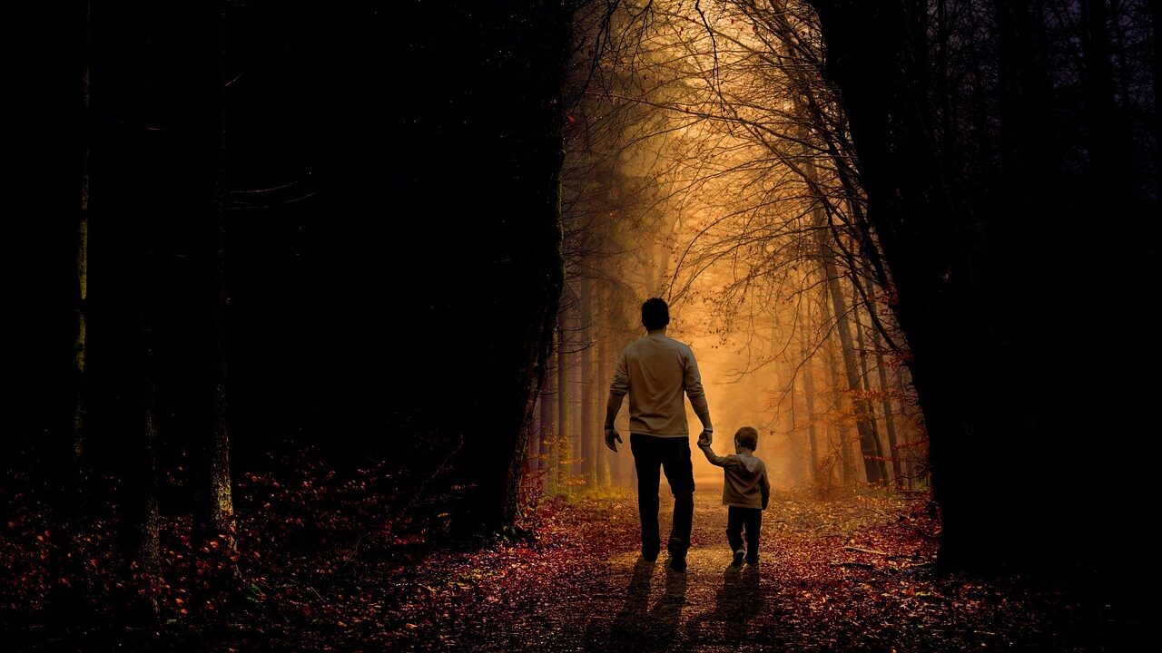 A father holds his young son's hand as they walk together down a forest path, symbolising the father-son bond and the healing of father wounds in counselling.