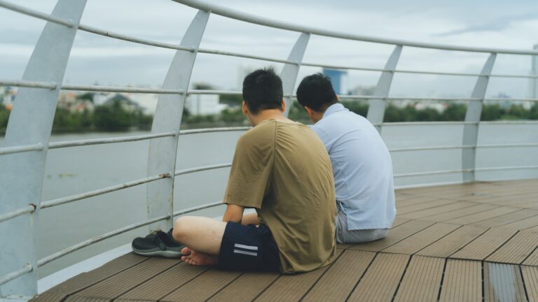TTwo men sitting side by side by the riverside — representing male friendship, emotional connection, and men's mental health support in Singapore