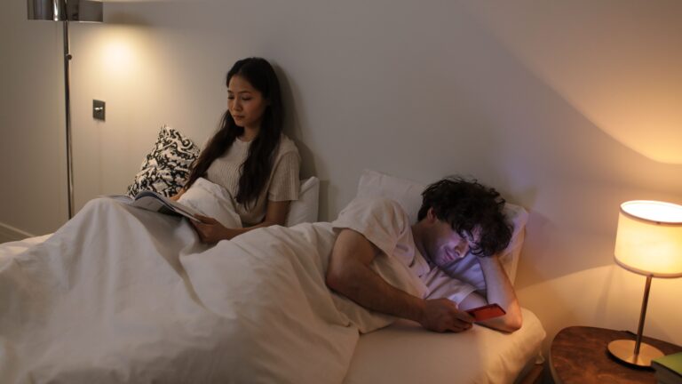 A couple lying in bed at night, physically close but emotionally distant. The woman sits upright reading a book, her expression quiet and withdrawn. The man lies beside her, face lit by the glow of his phone screen, turned away. The image illustrates the silent disconnection that pornography use can create in a marriage — two people sharing the same bed but no longer truly present with each other.