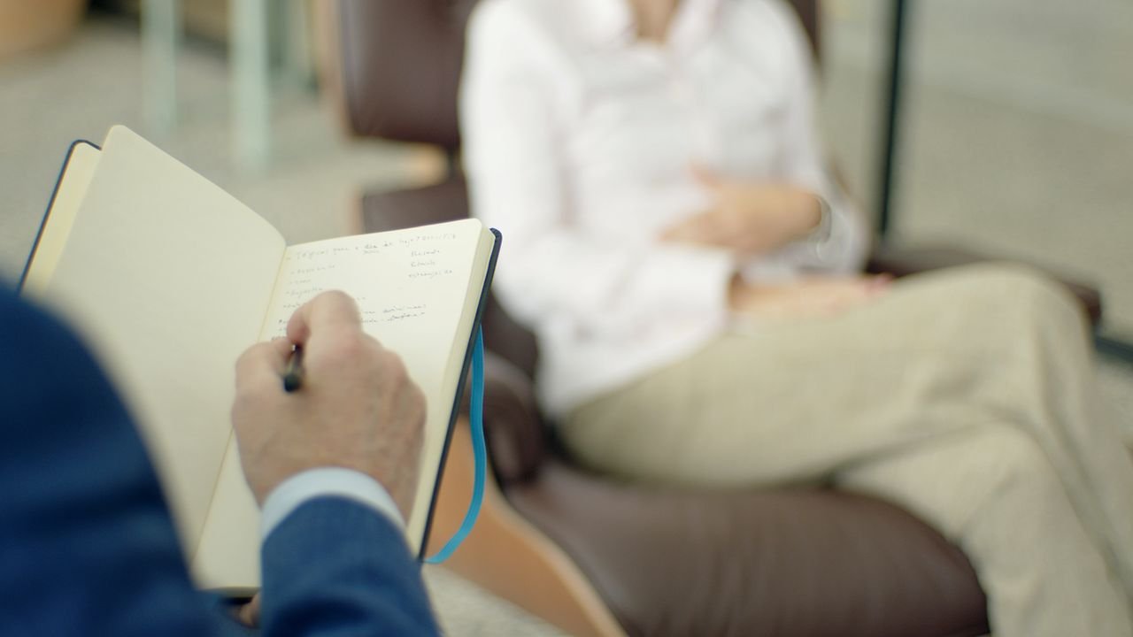 Close-up of a counsellor's hand writing notes in a notebook while a client sits across from them in a counselling session.