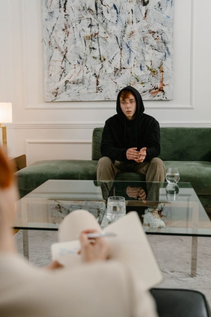 pexels-photo-7699484-7699484 A man in a black hoodie sits on a couch, attending a therapy session indoors.