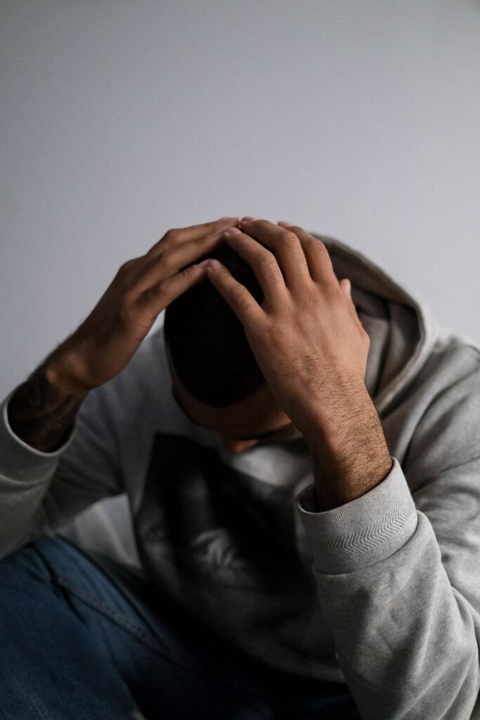 A man in a hoodie sits indoors, appearing contemplative or sad, with a focus on his hands and posture.